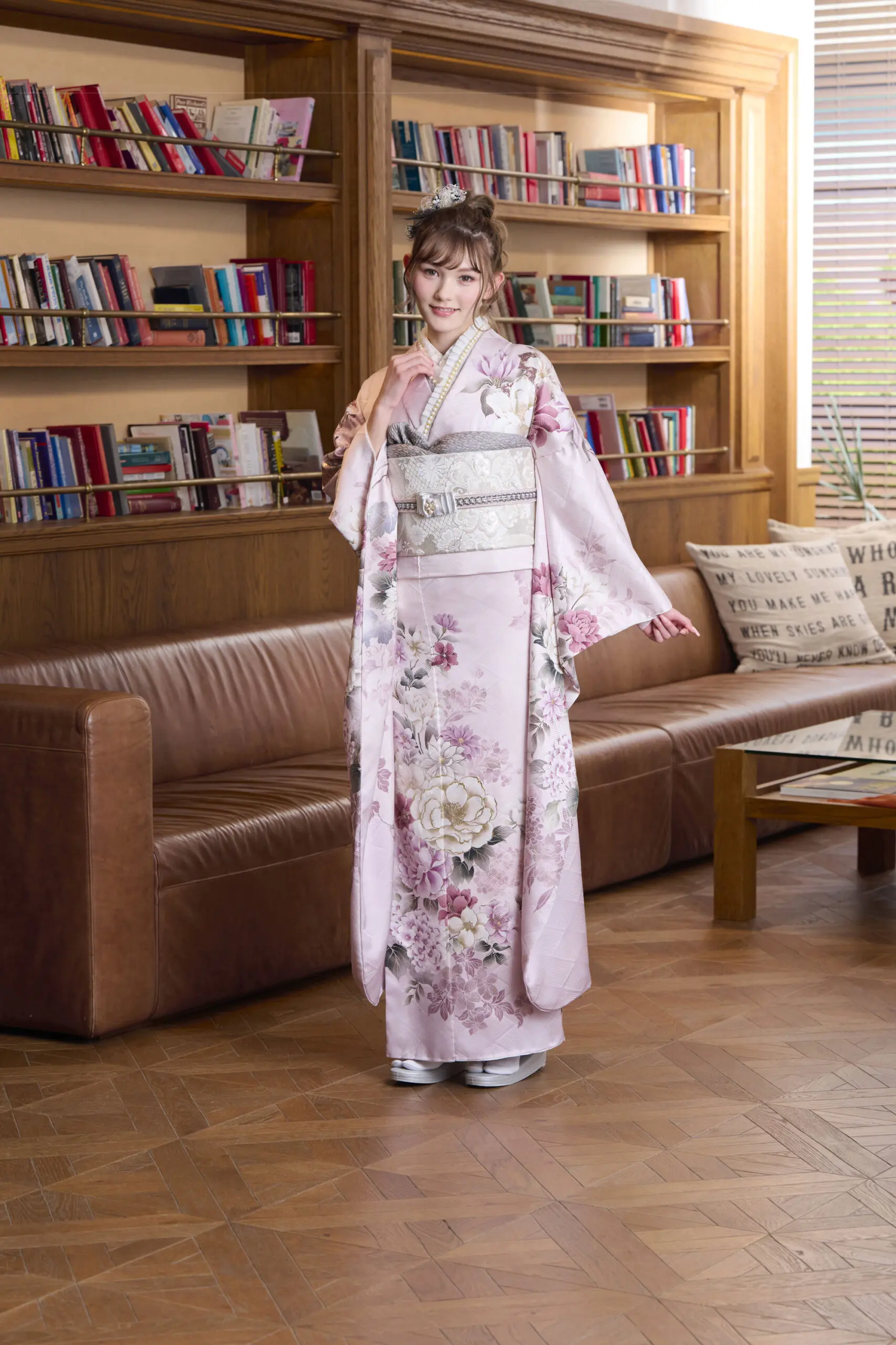 M-406の薄ピンクの花柄の着物を着た人が、木製の本棚、茶色の革張りのソファ、大きな文字がプリントされたクッション、フローリングの床、ブラインドのある窓のある部屋に立っている。.