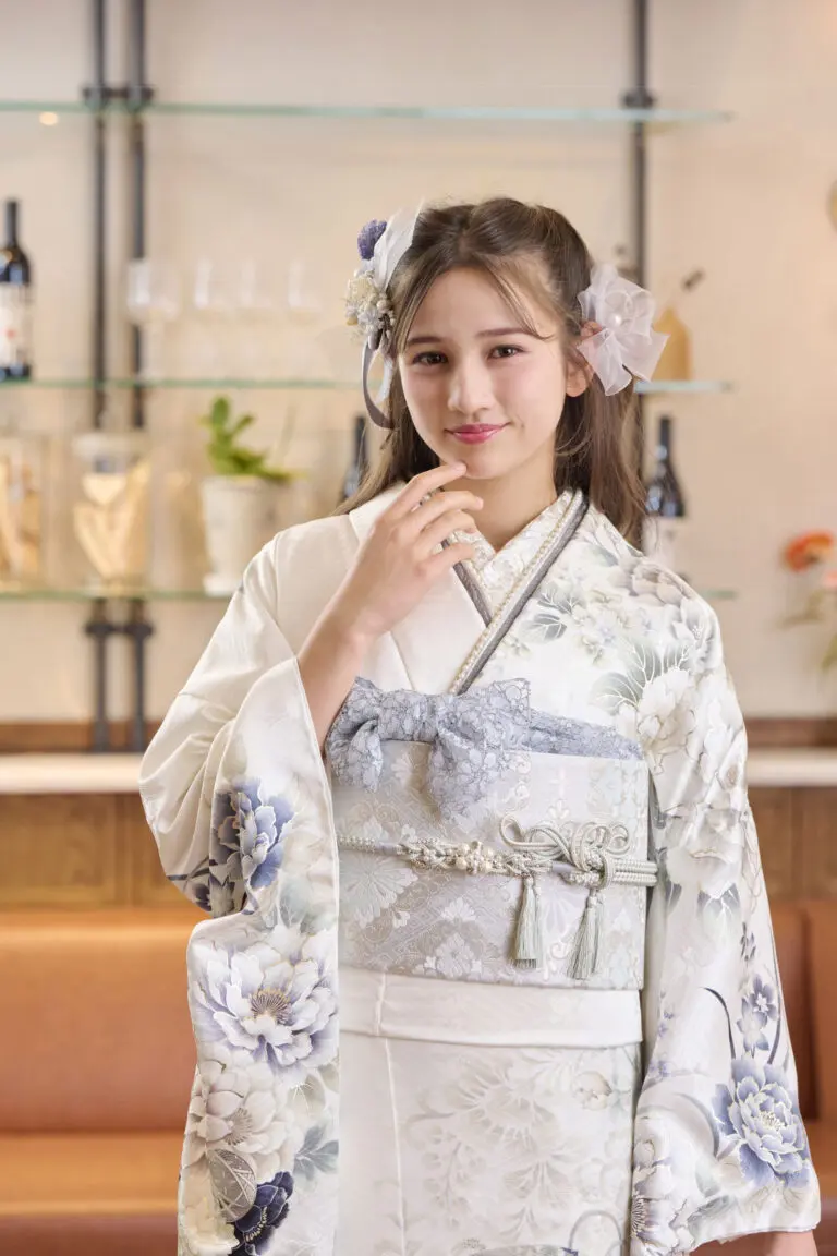 白と水色の花柄の着物を着たM-410のモデルを務める女性が、室内でボトルやガラス製品が並ぶガラス棚の前に立ち、片手を顔の近くに上げて柔らかく微笑んでいる。.