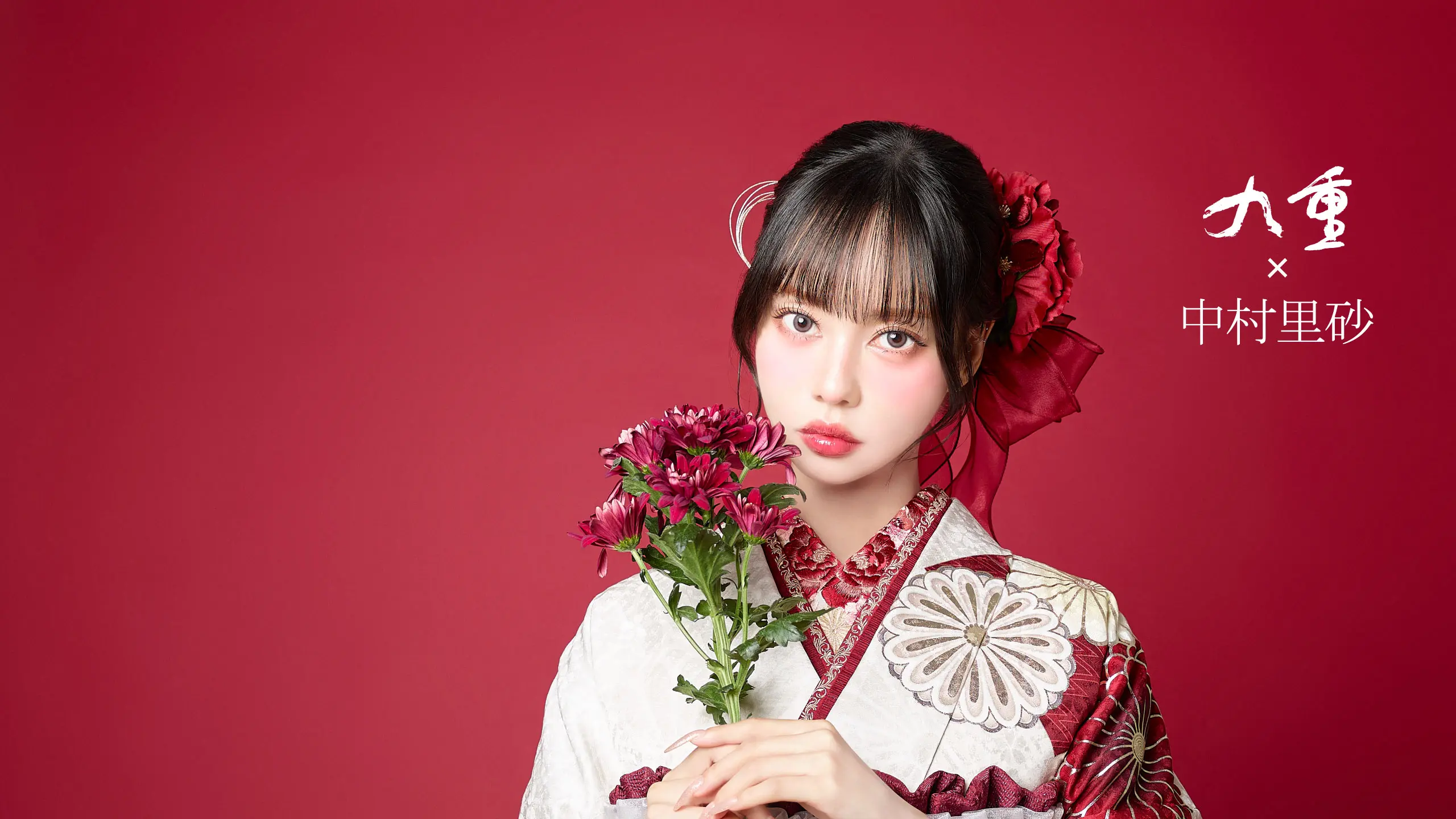柄物の着物を着た若い女性が、赤無地を背景にえんじ色の花束を持っている。前髪のあるストレートの黒髪に大きな赤いヘアアクセサリー。右上に日本語のテキストがある。.