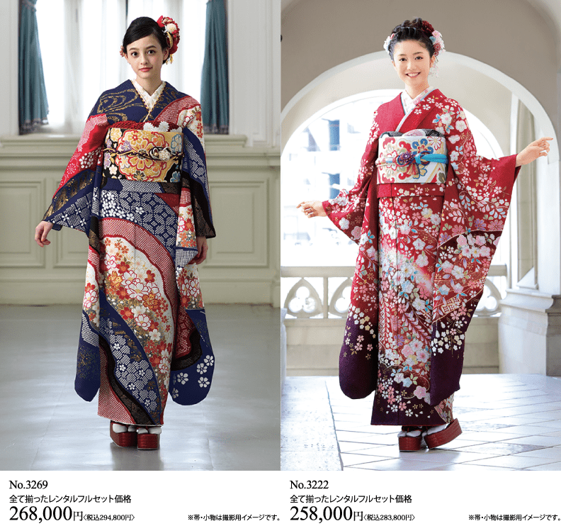 花柄の日本の伝統的な着物を着た2人の女性が室内に立っている。左の女性は紺色の着物を、右の女性は赤い着物を着ている。二人とも髪に花をつけて微笑んでいる。.