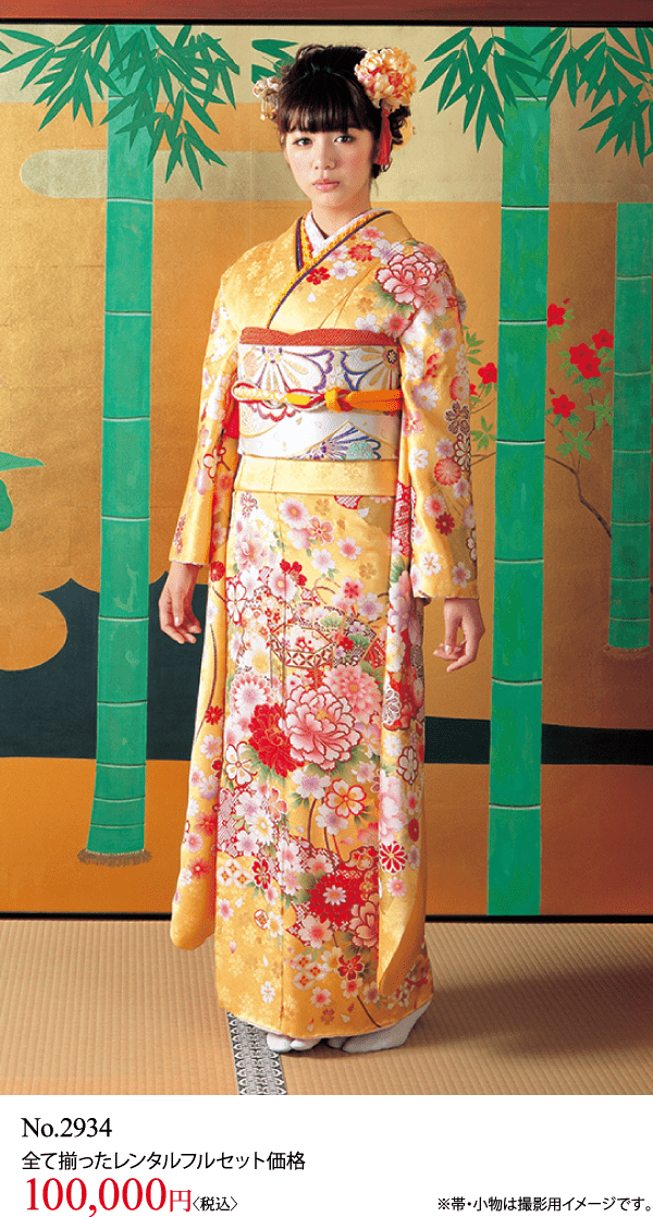 黄色とピンクの花柄の伝統的な着物に帯を締め、竹と木で飾られた金と緑の屏風の前に立つ女性。髪に花を飾り、カメラを見つめている。.