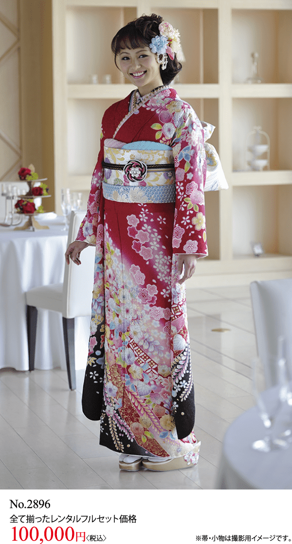 赤い花柄の着物に飾り帯をした女性が室内で微笑みながら立っている。花柄のヘアアクセサリーをつけている。背景には白いリネンのテーブルと椅子。.