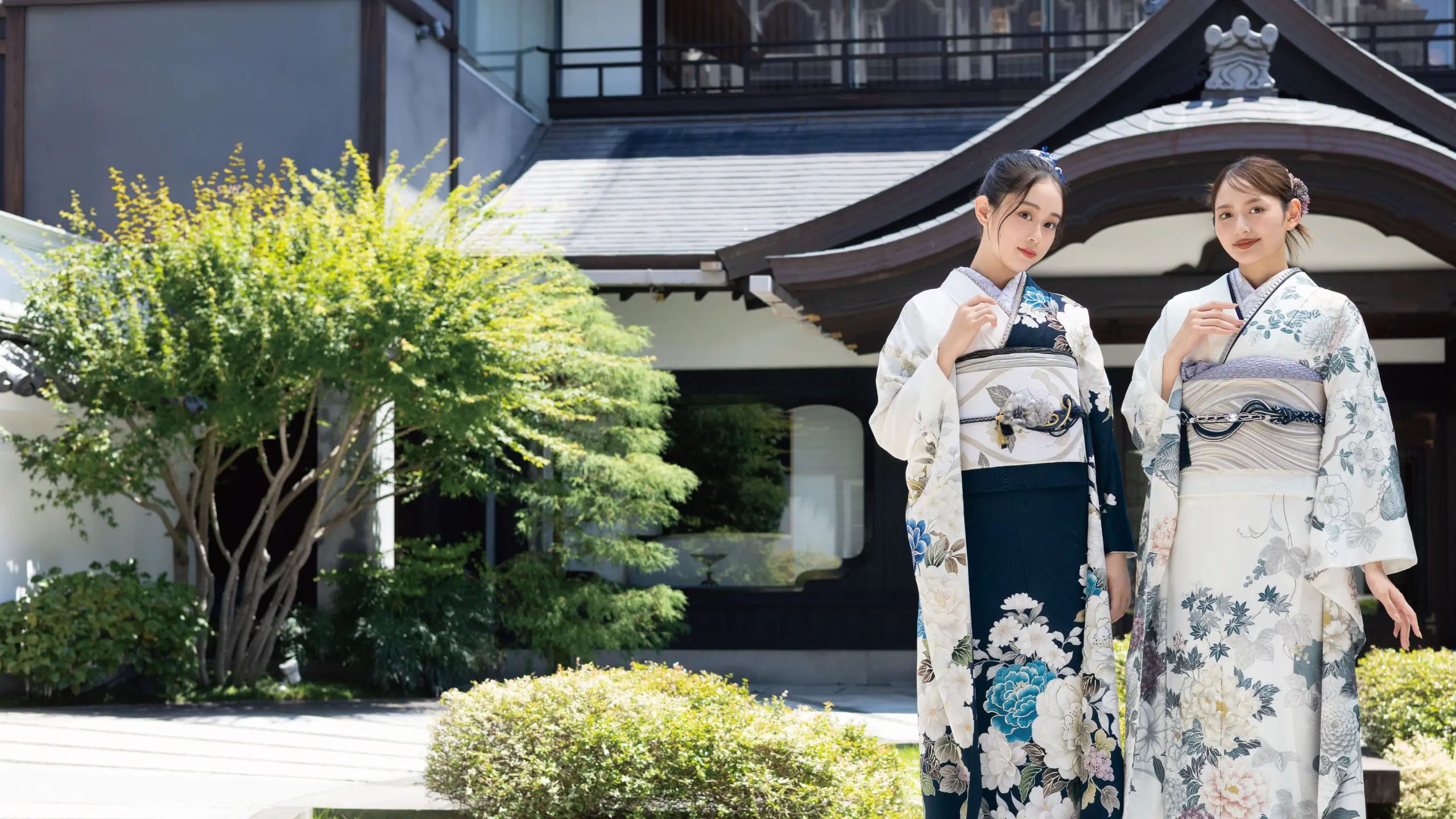 日本の伝統的な着物を着た2人の女性が、湾曲した屋根の建物の外で扇子を持って立っている。手前には緑の低木と木が見える。.
