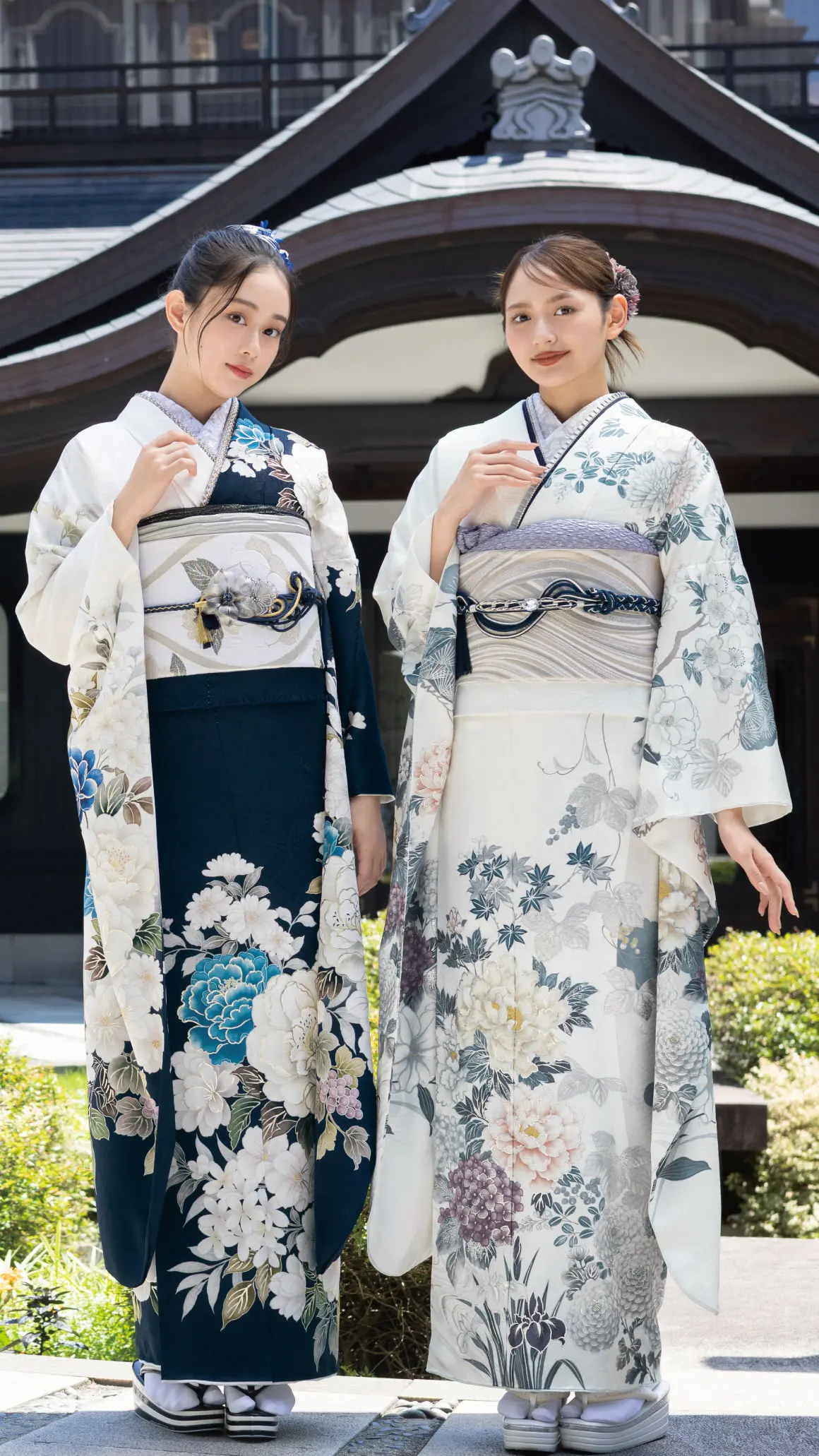 日本の伝統的な建物の前で、帯を締めたエレガントな花柄の着物を着た2人の女性が屋外に立っている。2人とも髪をきれいに整え、カメラの方を見ている。.