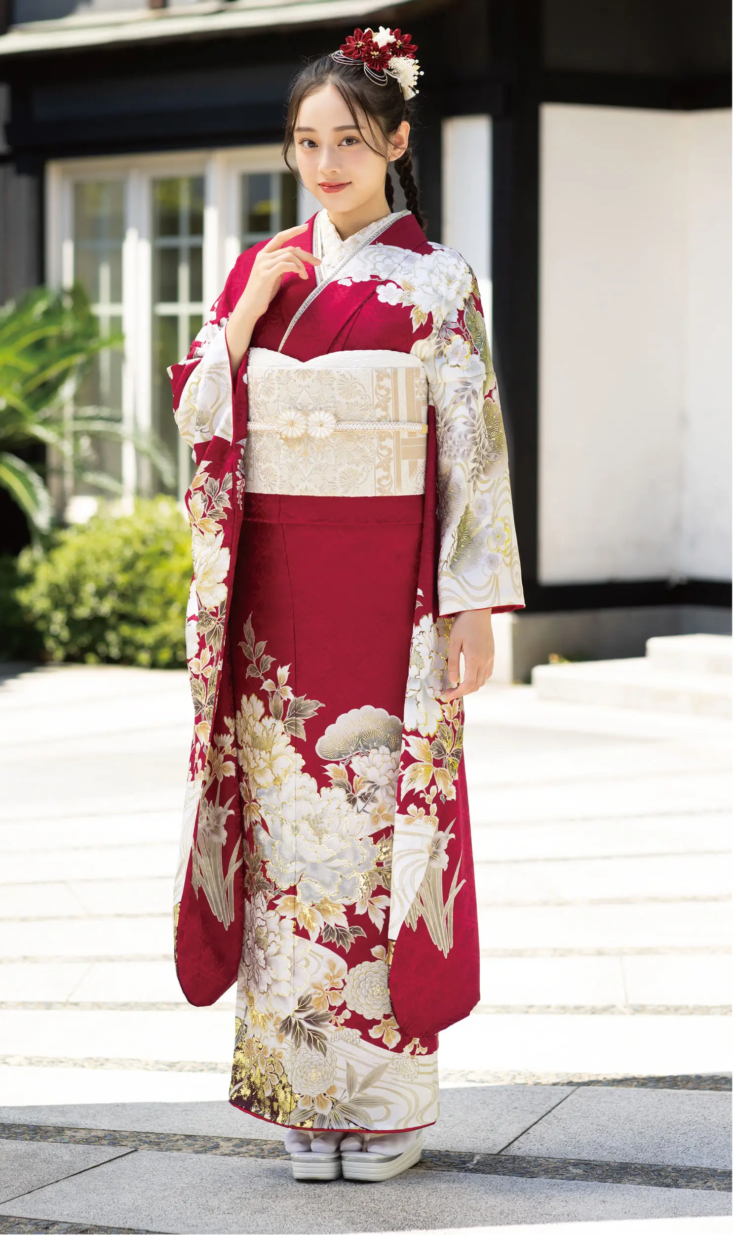 屋外で、K101の赤と白の花柄の着物にベージュの帯、花の髪飾り、伝統的な草履を合わせた女性。背景は緑と建物。.