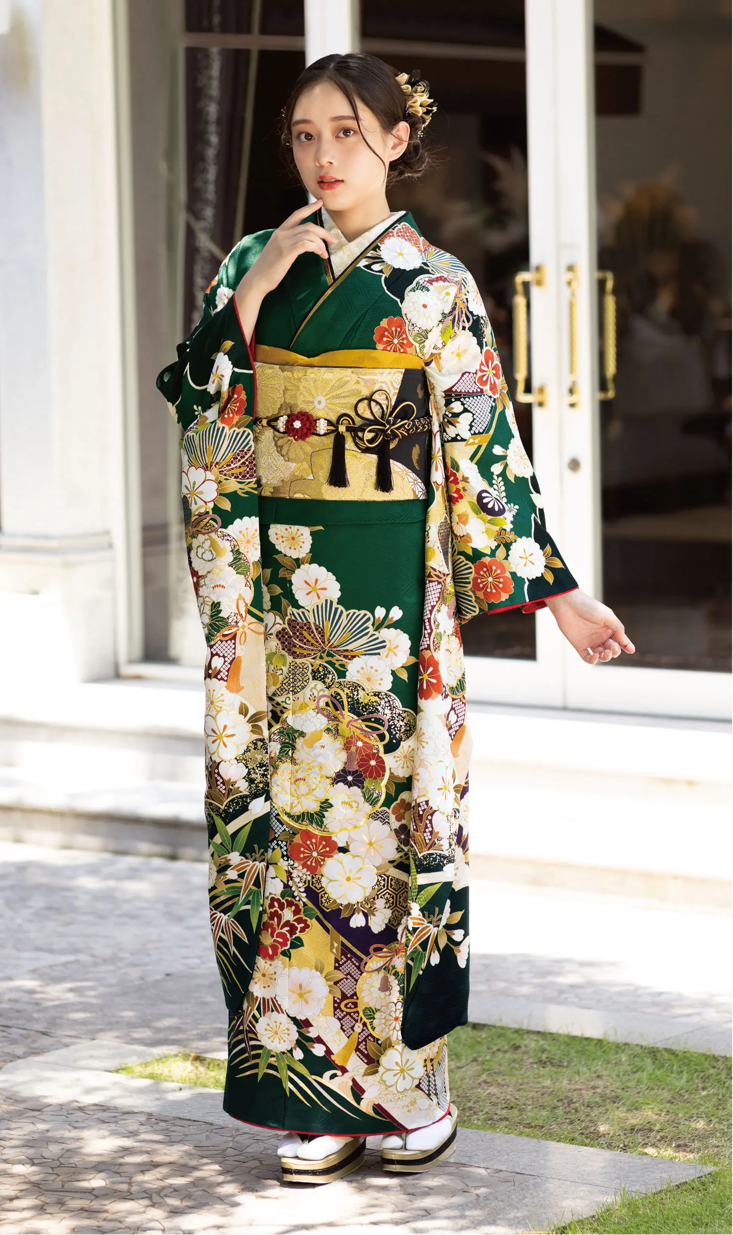 ダークグリーンとゴールドの花柄プリントのK101着物にゴールドの帯を締め、ガラス戸の前に立つ女性。髪はかんざしでアップにし、伝統的な草履に白い靴下を合わせている。.