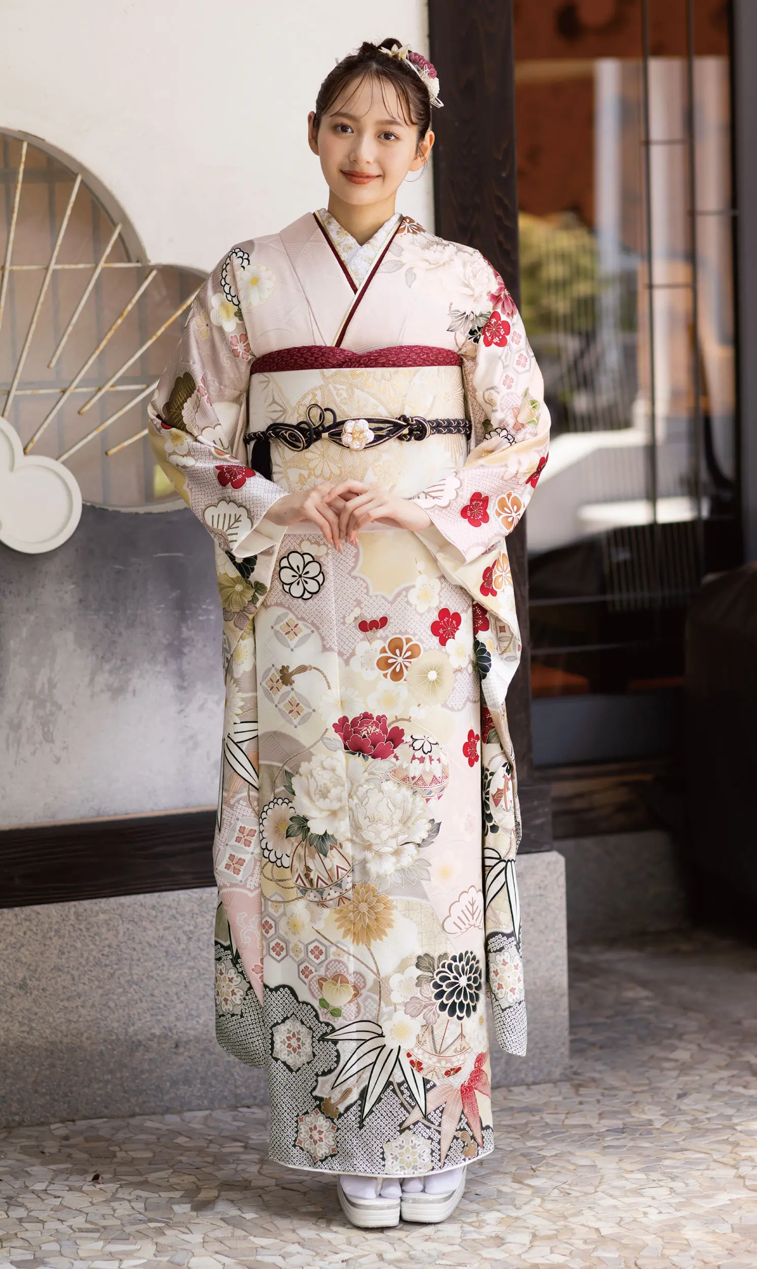 花柄の淡い色の着物に帯を締め、草履を履いた若い女性。髪をきれいに整え、穏やかな笑顔で前を向いている。.