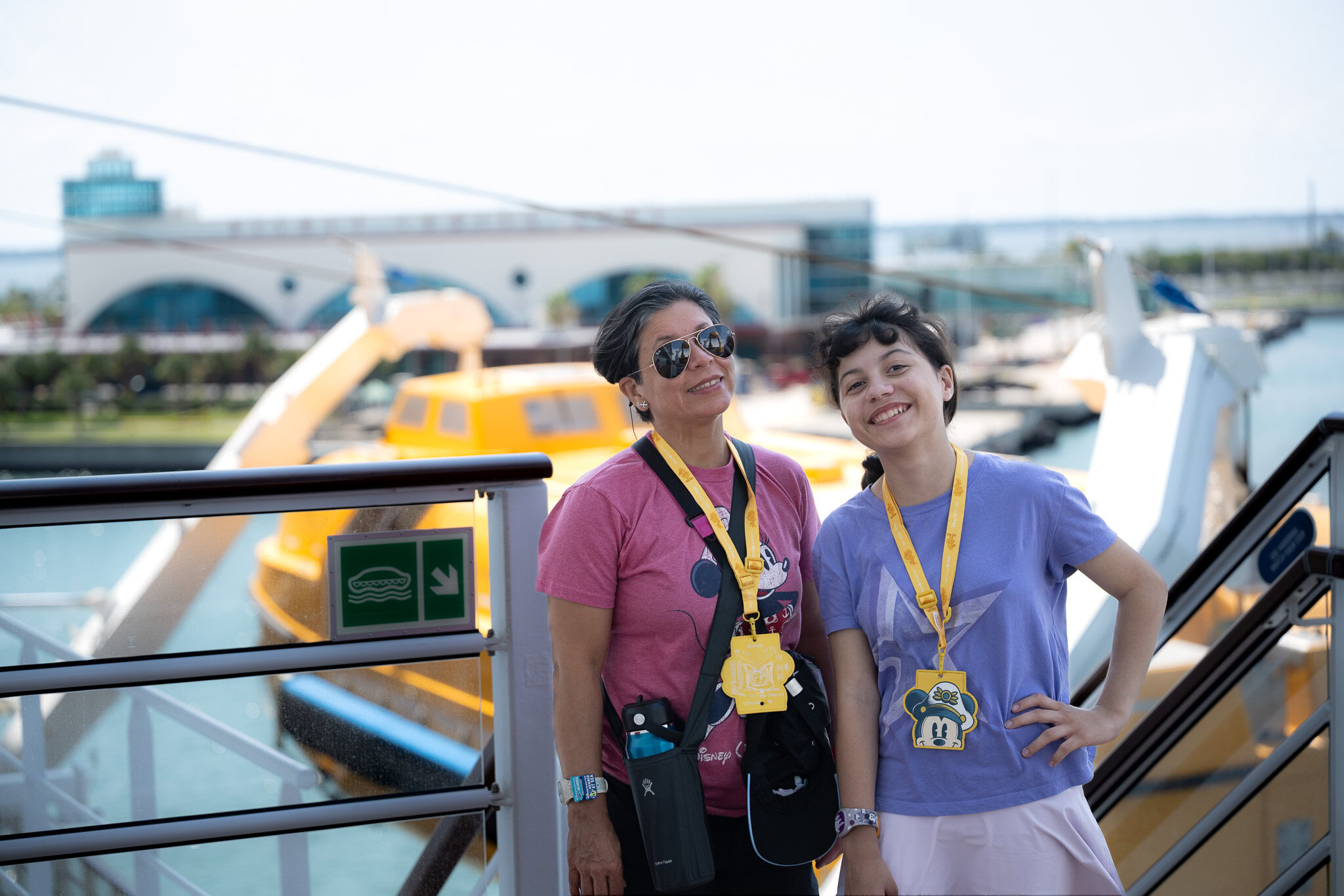 Lucy and Daniela from Laugh and Gear smiling aboard the Disney Wish cruise ship at Port Canaveral, wearing Disney lanyards, with the cruise terminal and a yellow tender boat in the background.