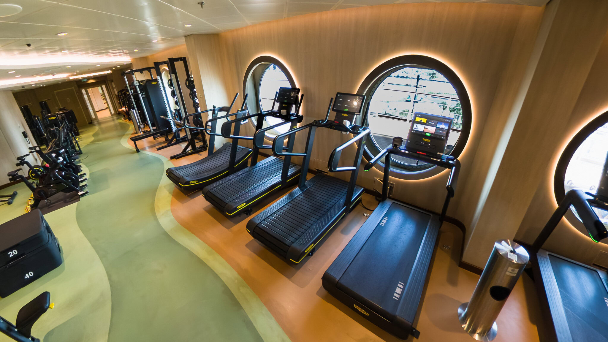 Modern fitness center on the Disney Wish featuring treadmills, weight machines, and large porthole windows overlooking the ocean.