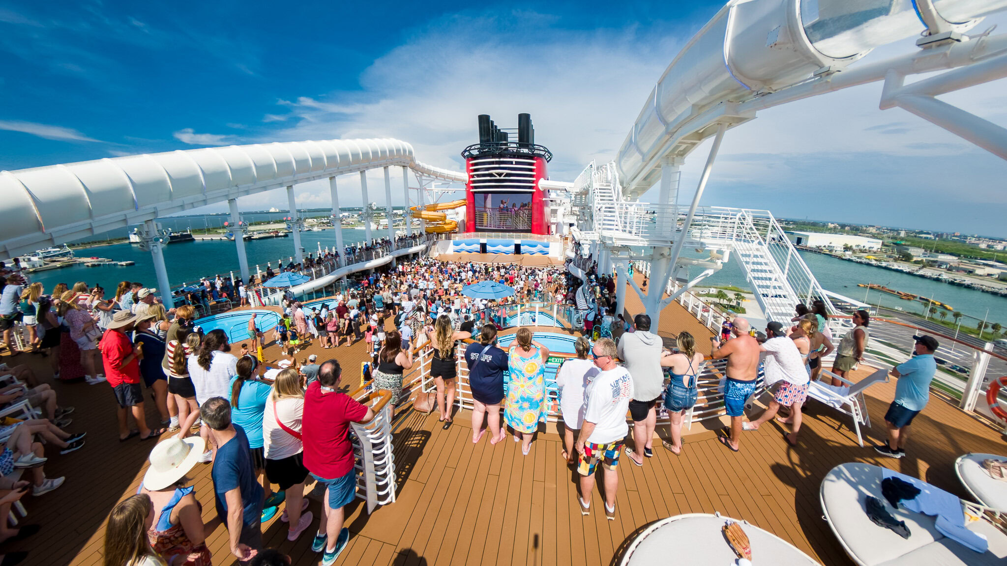 Crowds gathered on the Disney Wish upper deck during the Sail Away Party with AquaMouse slides and the ship’s funnel in the background.