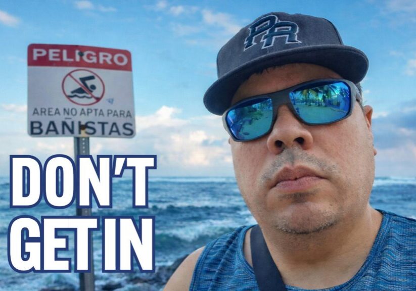 Man standing near rough ocean waves in San Juan next to a “PELIGRO” sign warning that the area is not safe for swimmers.