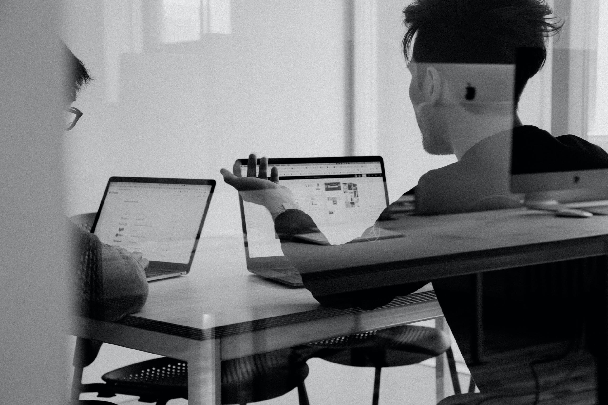 Duas pessoas estão sentadas em uma mesa, trabalhando em seus laptops. Uma pessoa está apontando para a tela e a outra está olhando para o monitor. Vocês dois estão em um ambiente de escritório moderno e provavelmente estão se preparando para uma reunião importante sobre sua estratégia DealShaker.