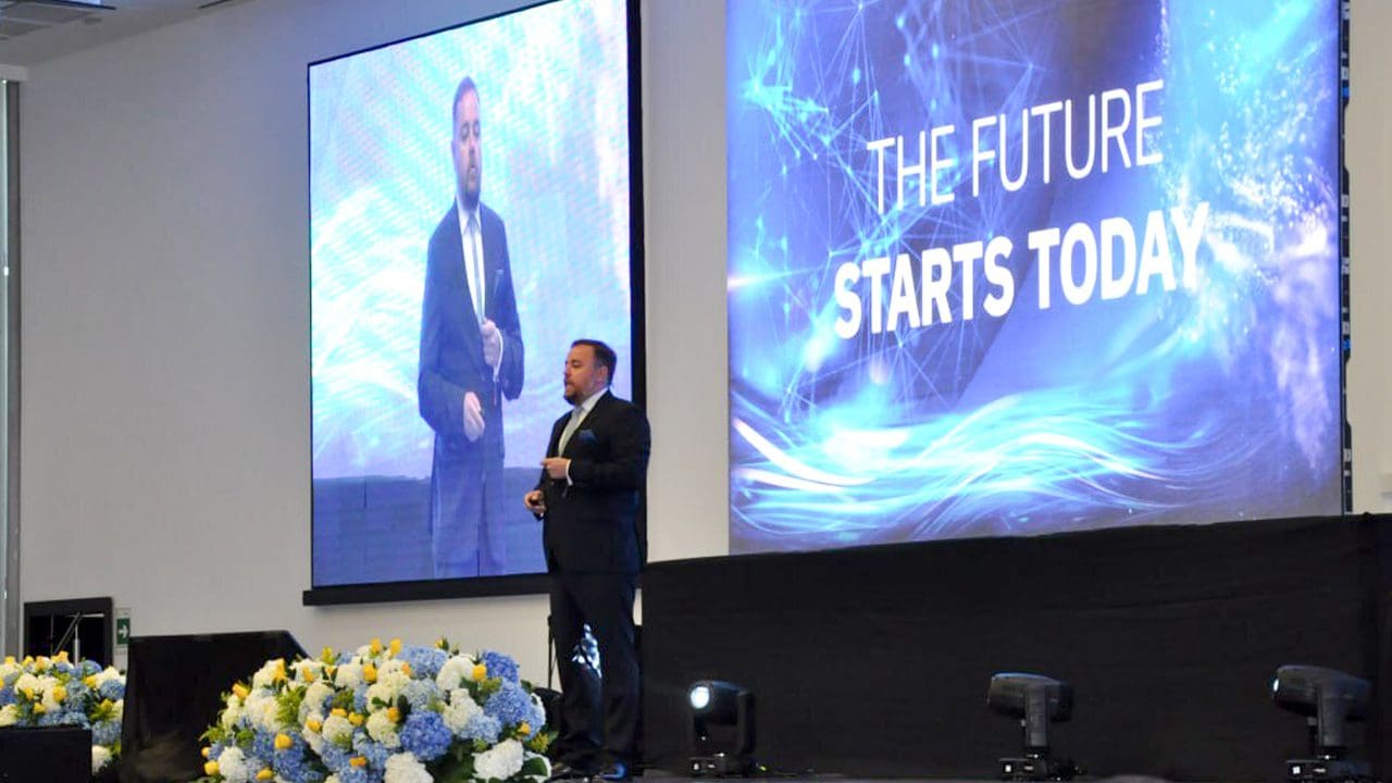 Un hombre de traje está de pie en un escenario junto a una gran pantalla. Las palabras "EL FUTURO COMIENZA HOY" se proyectan en la pantalla. Parece estar dando una presentación en un evento corporativo de OES. El escenario está decorado con flores.