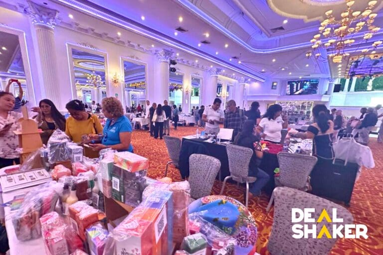 Di ruangan yang cerah dan berornamen yang dipenuhi dengan kolom yang dihias, orang-orang melihat-lihat dan berinteraksi dengan berbagai stan. DI SUDUT KANAN BAWAH, ANDA DAPAT MELIHAT LOGO "DEALSHAKER", MENYOROTI KEMITRAAN DENGAN BERBAGAI VENDOR PEDAGANG YANG MENERIMA SATU CRYPTOCURRENCY UNTUK PENJUALAN BARANG DAGANGAN.