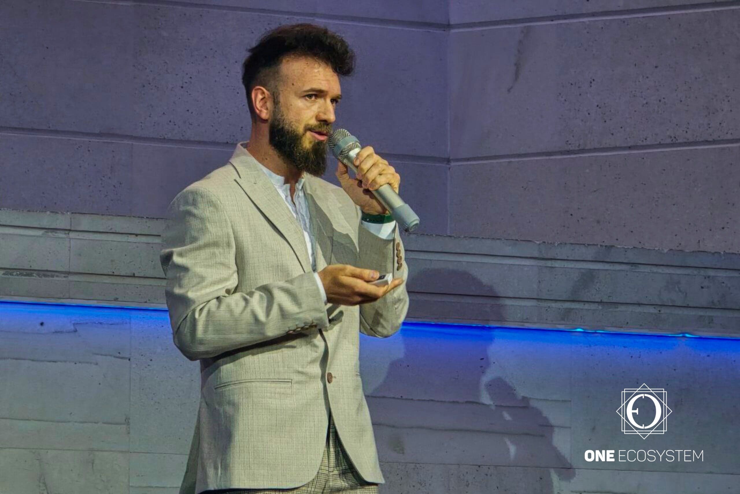 Një burrë me mjekër dhe i zbukuruar mirë po flet në një mikrofon ndërsa mban një pajisje të vogël në sfondin e një muri guri me vija blu dhe një logo që thotë "NJË EKOSISTEM" në qoshe. I shquari Ivan Zerkov ofron njohuri në lidhje me blerjet e mysafirëve dhe tregtarët.