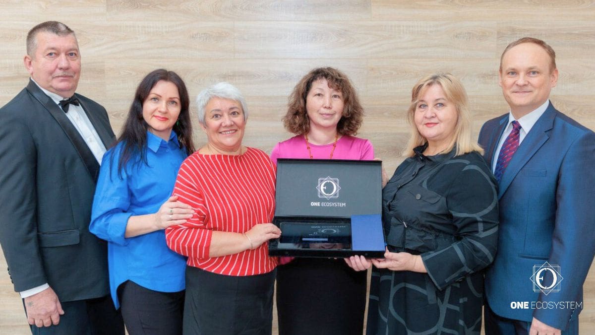 Um grupo de 6 pessoas, 3 homens e 3 mulheres, em pé dentro de um evento da IMA em Novosibirsk. Eles estão vestidos com trajes formais, segurando laptops com "One Ecosystem" exibido na tela e sorrindo para a câmera.