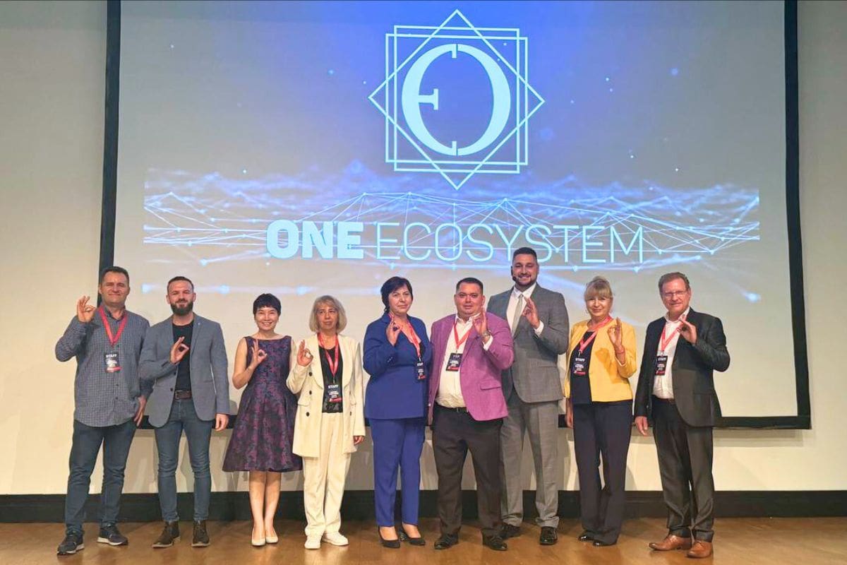 무대에는 9명이 줄을 서 있고, 배경에는 'One Ecosystem' 로고가 빛나며 손을 흔든다. 이 장면은 IMA 이벤트의 일부로, 흥미로운 새 기능을 공개하고 루마니아의 혁신적인 정신을 전 세계 청중에게 전달합니다.