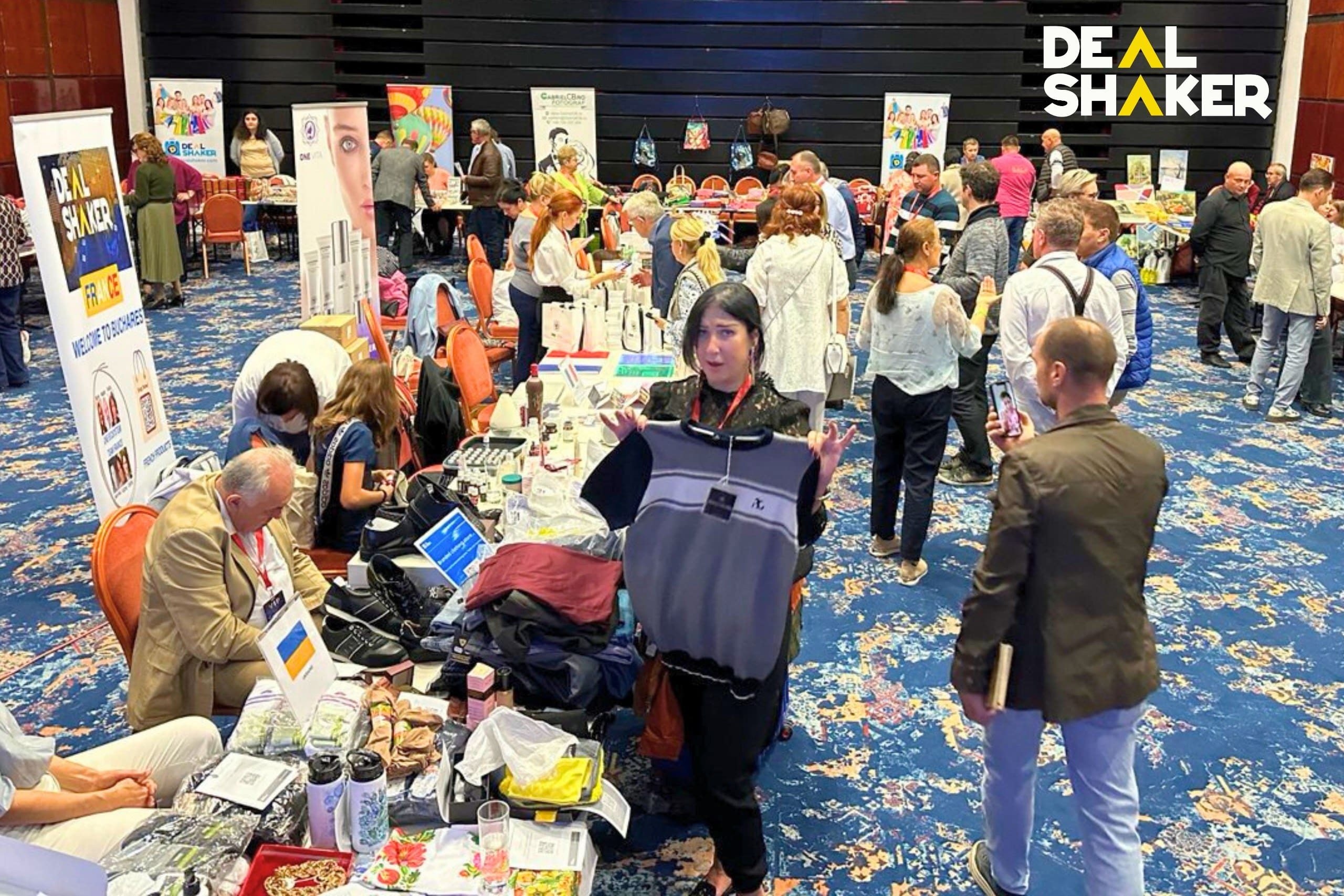 Pasar dalam ruangan yang semarak menyambut pengunjung, dan pembeli yang bersemangat menelusuri berbagai barang di berbagai kios. Logo "Deal Shaker" berdiri dengan bangga di dinding, mengisyaratkan kehadirannya yang semarak di Deal Shaker Expo. Semua ini terungkap dalam suasana hidup yang mengingatkan pada bazar Rumania.