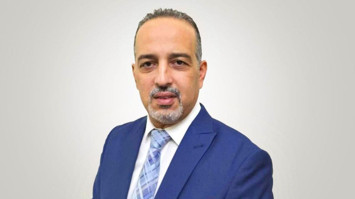 Un bărbat într-un costum albastru și cravată cu model face o prezentare încrezătoare în fața camerei pe un fundal neutru, ca și cum ar fi la o conferință internațională a consiliului de administrație al țării.