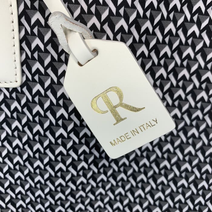 白と黒の幾何学模様の布地を背景に、ゴールドの「R」の文字と「MADE IN ITALY」の文字が入った白い革製のROBERTA PIERIタグがぶら下がっている。.