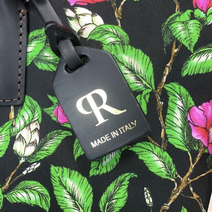 ゴールドの「R」とイタリア製テキストをあしらったブラックのROBERTA PIERIラゲッジタグが、グリーンの葉とピンクの花をあしらった花柄のファブリックに吊り下げられ、ミニダッフルを時代を超えたイタリアンエレガンスで引き立てます。.