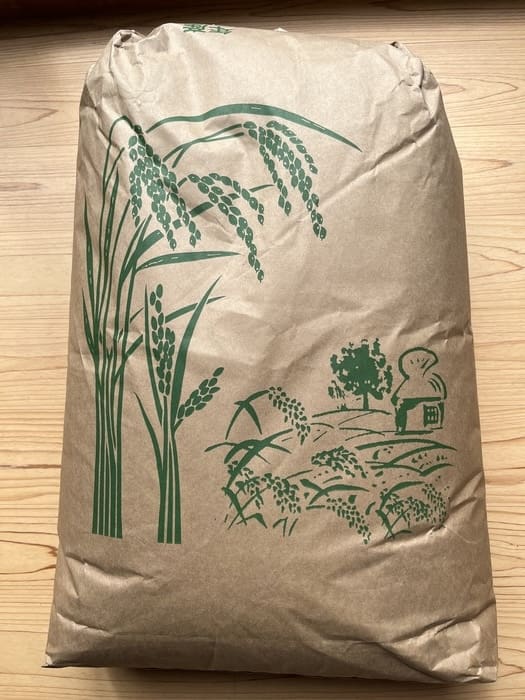稲と田園風景の緑のイラストが描かれた大きな茶色の紙袋が、信濃町産米コシヒカリをアピールしている。明るい木の上に置かれている。.