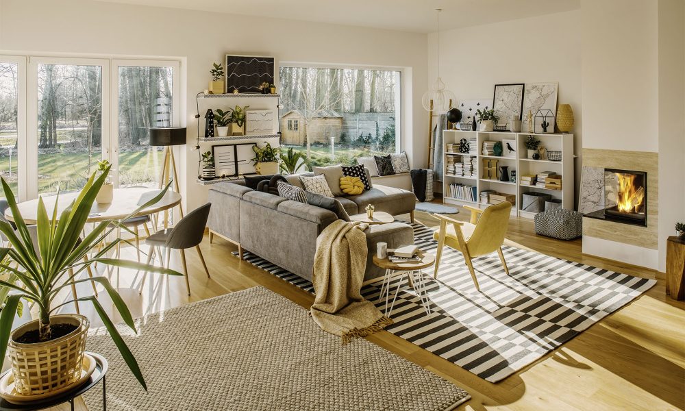 Two carpets on the floor in white Scandi living room interior with big window, glass door, corner sofa, fireplace and fresh plants
