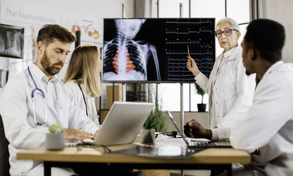 Caucasian aged woman in white lab coat standing near big monitor and ponting on heartbeat rates during briefing with competent international doctors. Concept of medicine and health care.