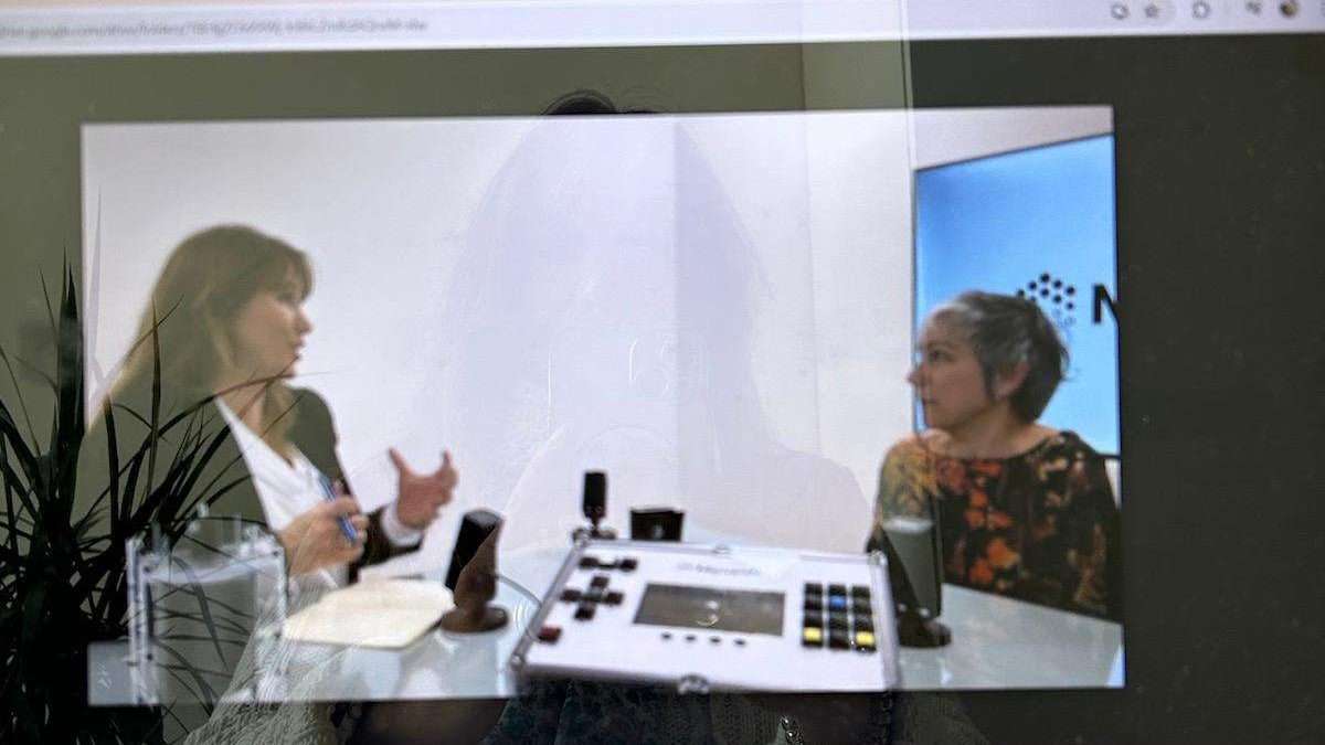 Duas mulheres sentadas em uma mesa discutem a transformação digital com microfones e equipamentos de áudio entre elas. A cena aparece em uma tela com um reflexo fraco de uma pessoa e uma planta em primeiro plano.