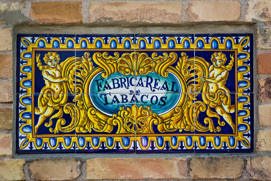 Azulejo de parede de tijolo com a inscrição "FÁBRICA REAL DE TABACOS" e decoração ornamental, ligada à Real Fábrica de Tabaco de Sevilha, identidade gráfica urbana.