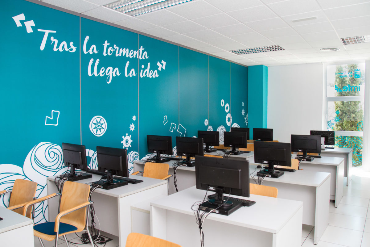 Sala de computadores do projeto IMFE com marcação espacial nas paredes turquesa, frases inspiradoras em grande formato e estações de computador distribuídas em filas.