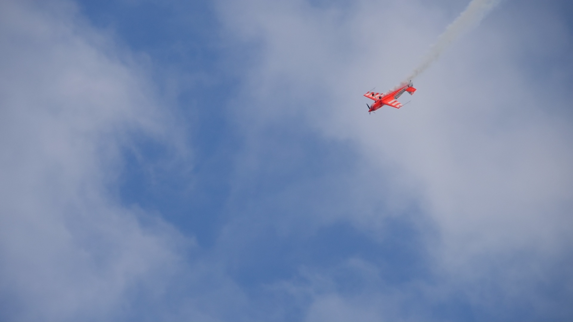 Un avión rojo de acrobacias vuela verticalmente hacia abajo a través de un cielo azul parcialmente nublado, dejando tras de sí una estela de humo blanco.