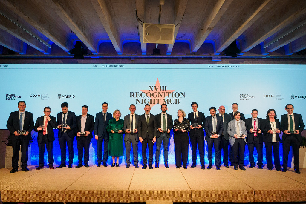 Un grupo de dieciséis personas en traje formal de pie en un escenario sosteniendo premios delante de un telón de fondo azul que lee XVIII NOCHE DE RECONOCIMIENTO MCB, celebrando los logros de los embajadores de congresos en Madrid.