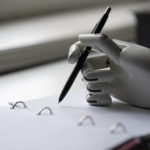 A robot's hand writing on a notepad with a pen.